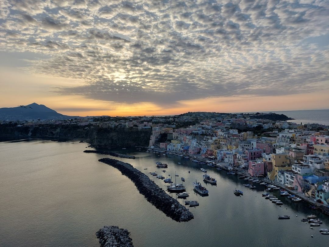 Sunset view over a colorful coastal village with boats anchored in a natural harbor, framed by dramatic clouds and a distant volcanic mountain.